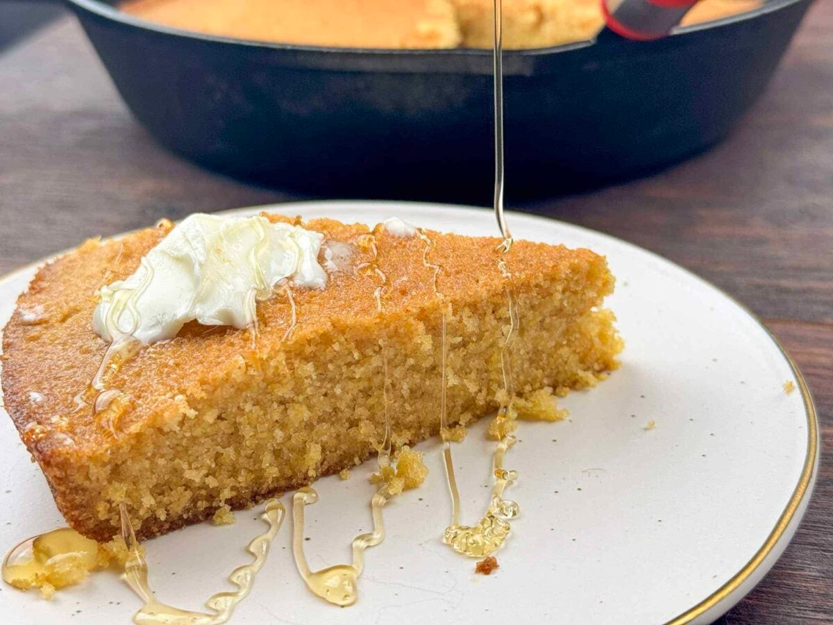 This is a slice of cornbread on a white plate with a drizzle of honey and pat of butter.