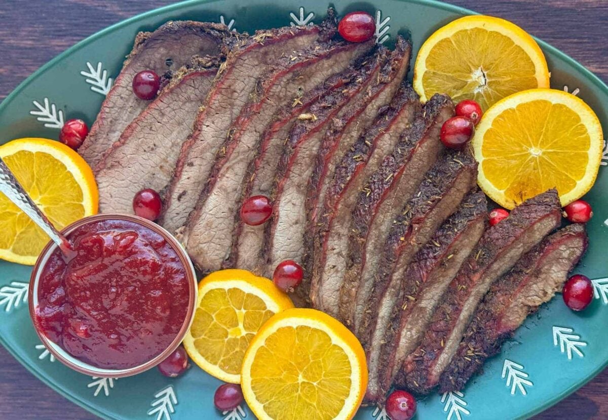 green christmas platter of sliced brisket with bowl of cranberry bbq sauce, slices of orange, fresh cranberry garnish.