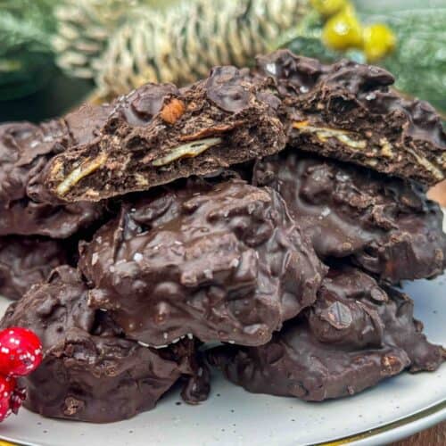 pile of crockpot christmas crack clusters on white plate sitting on wooden table with christmas foliage.