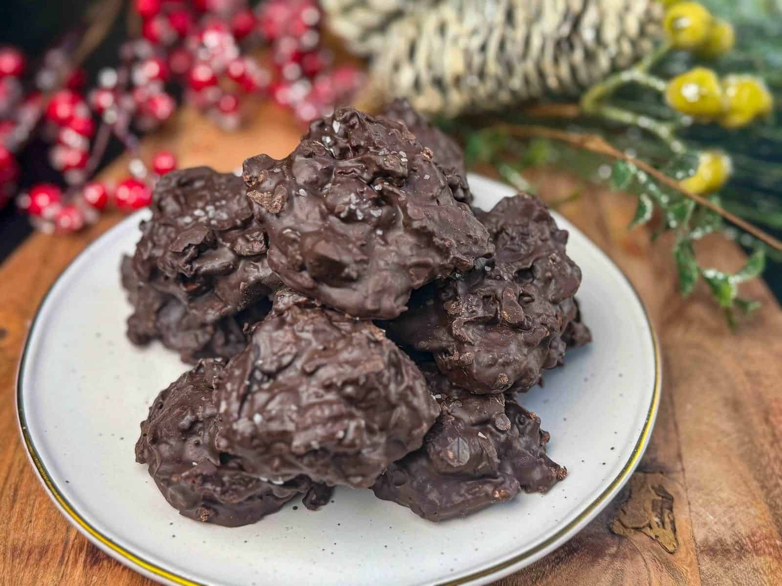 pile of crockpot christmas crack clusters on white plate sitting on wooden table with christmas foliage.
