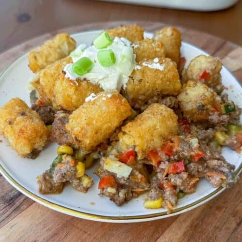 white plate of serving of healthy tater tot casserole topped with sourcream and green onion on wooden cutting board