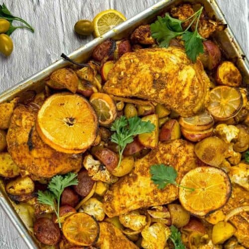 overhead angled view of the moroccan chicken sheet pan with garnish of lemon slices, parsley, and olives on gray backdrop