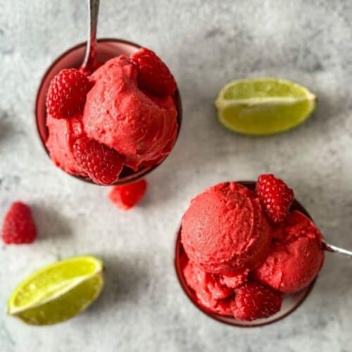 This is two small dishes with scoops of raspberry sorbet with spoons and lime wedges in the background.