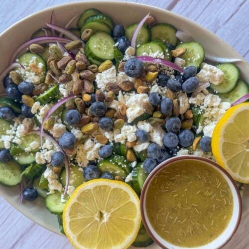 This is a platter of assembled cucumber blueberry salad with a dish of lemon vinaigrette and sliced lemon on the side of the platter.