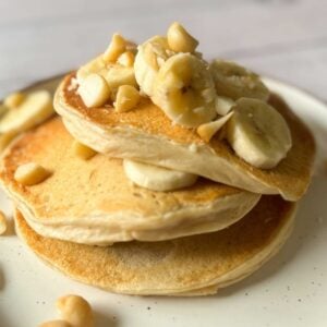 A stack of three pancakes on a plate topped with banana and macadamia nuts.