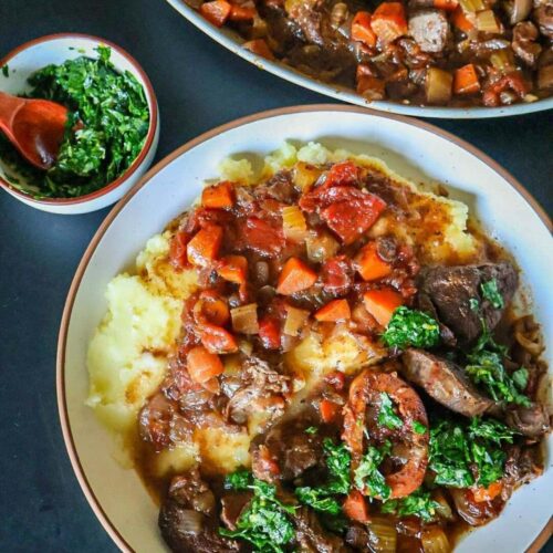 A while plate with bison osso buco served with mashed potatoes. A small dish of gremolata and the serving platter.