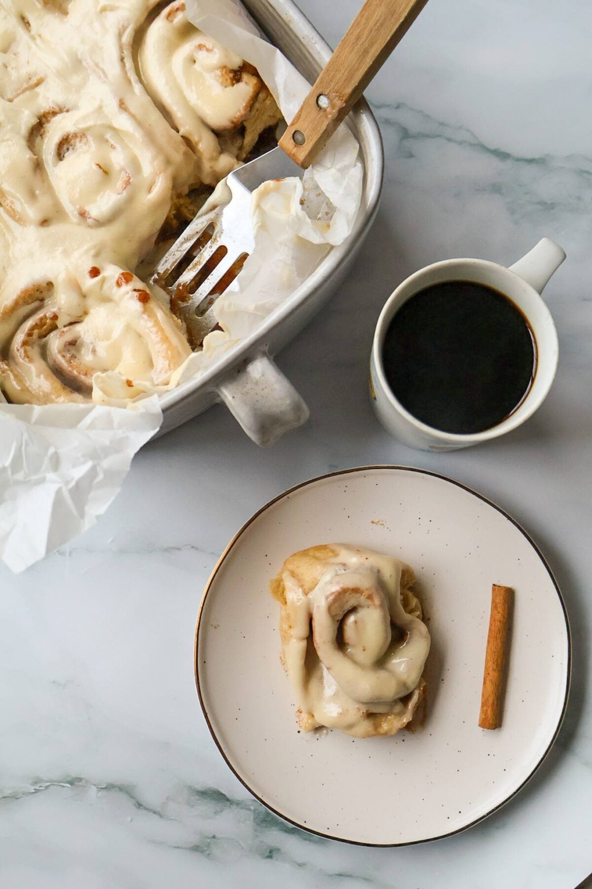 A white plate with a cinnamon roll, cinnamon stick, cup of coffee, next to the baking dish on marble backdrop.