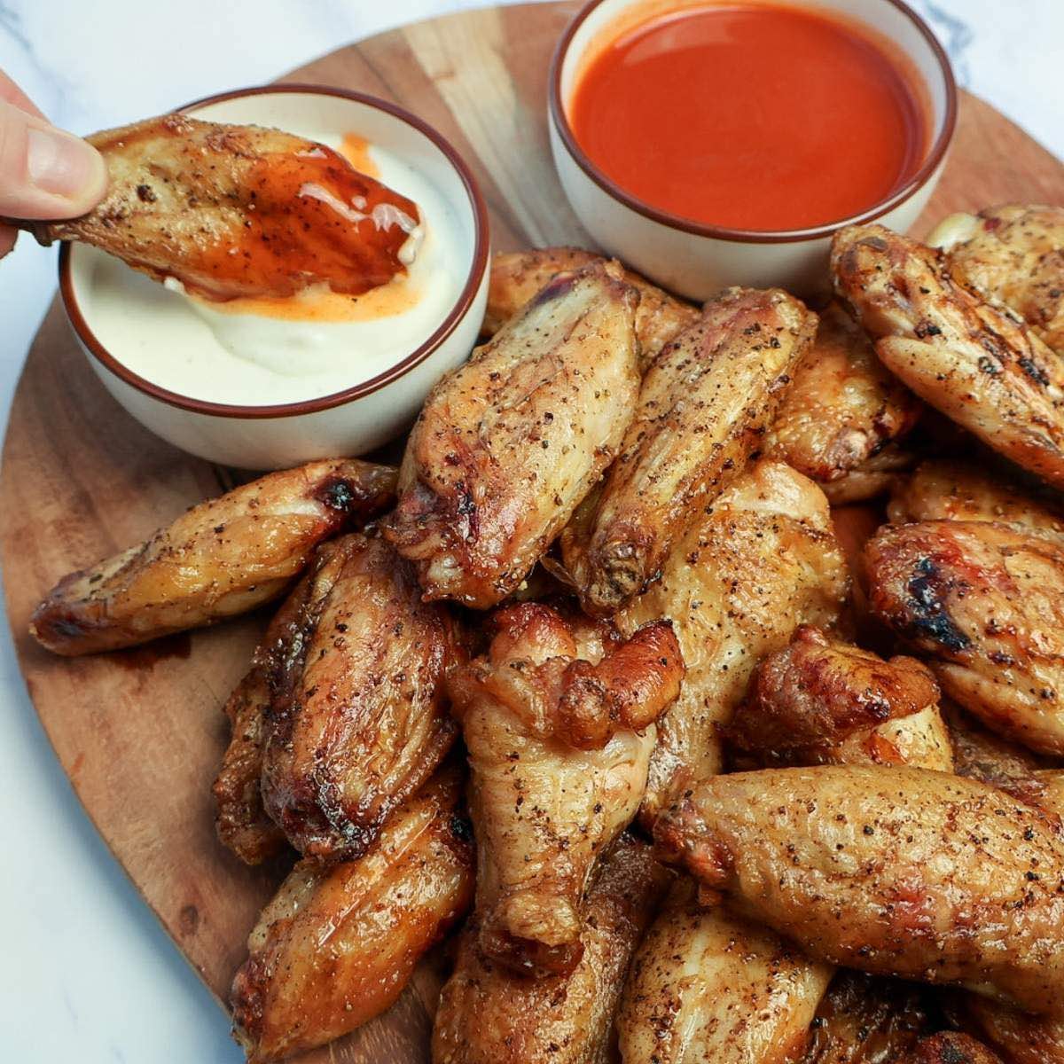 A platter of smoked wings with one being dipped in buffalo and ranch sauces.