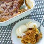 White plate of apple cake with vanilla ice cream with whole pan behind.