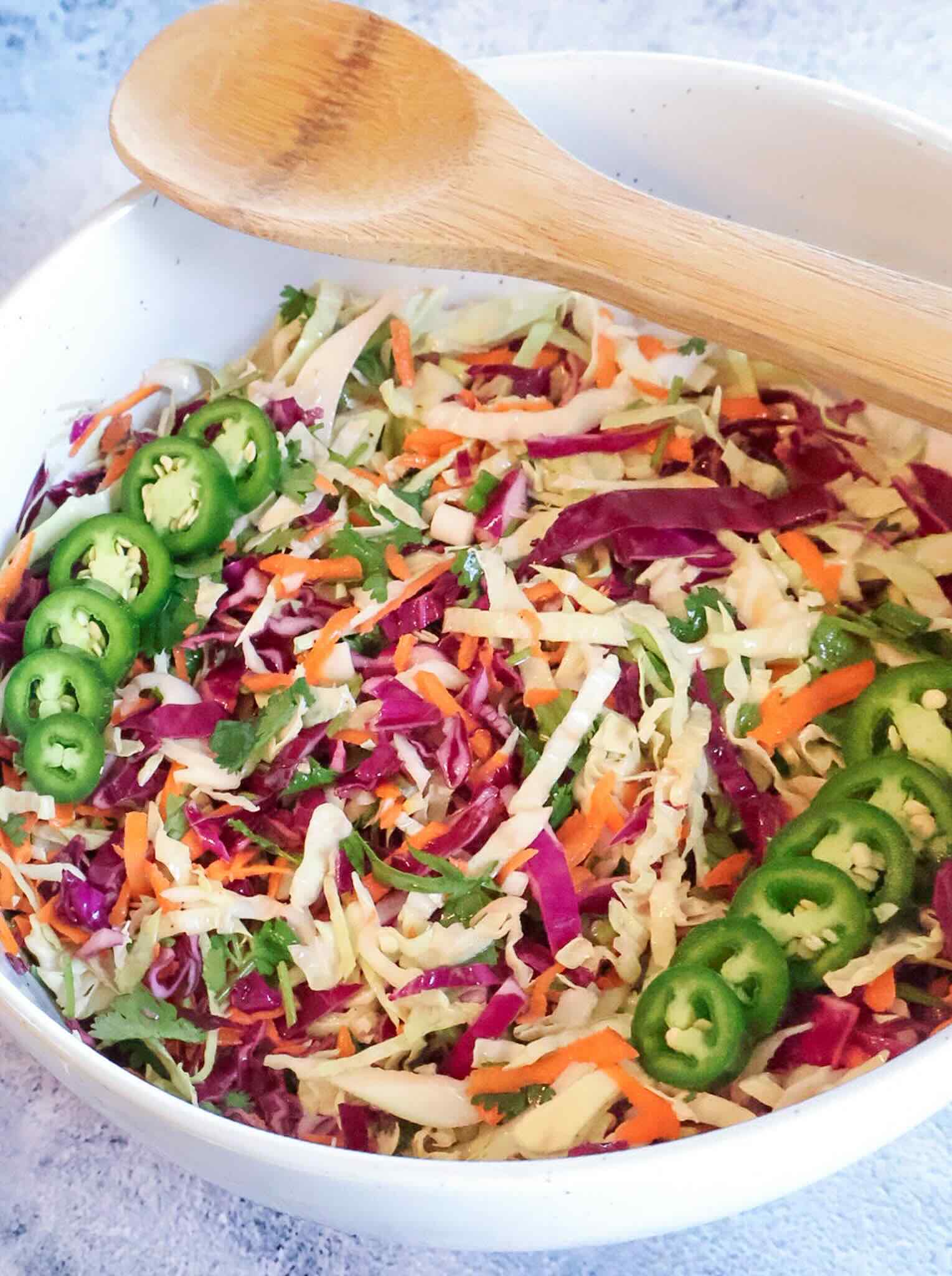 A white bowl of jalapeño lime slaw with a wooden spoon resting on the edge.