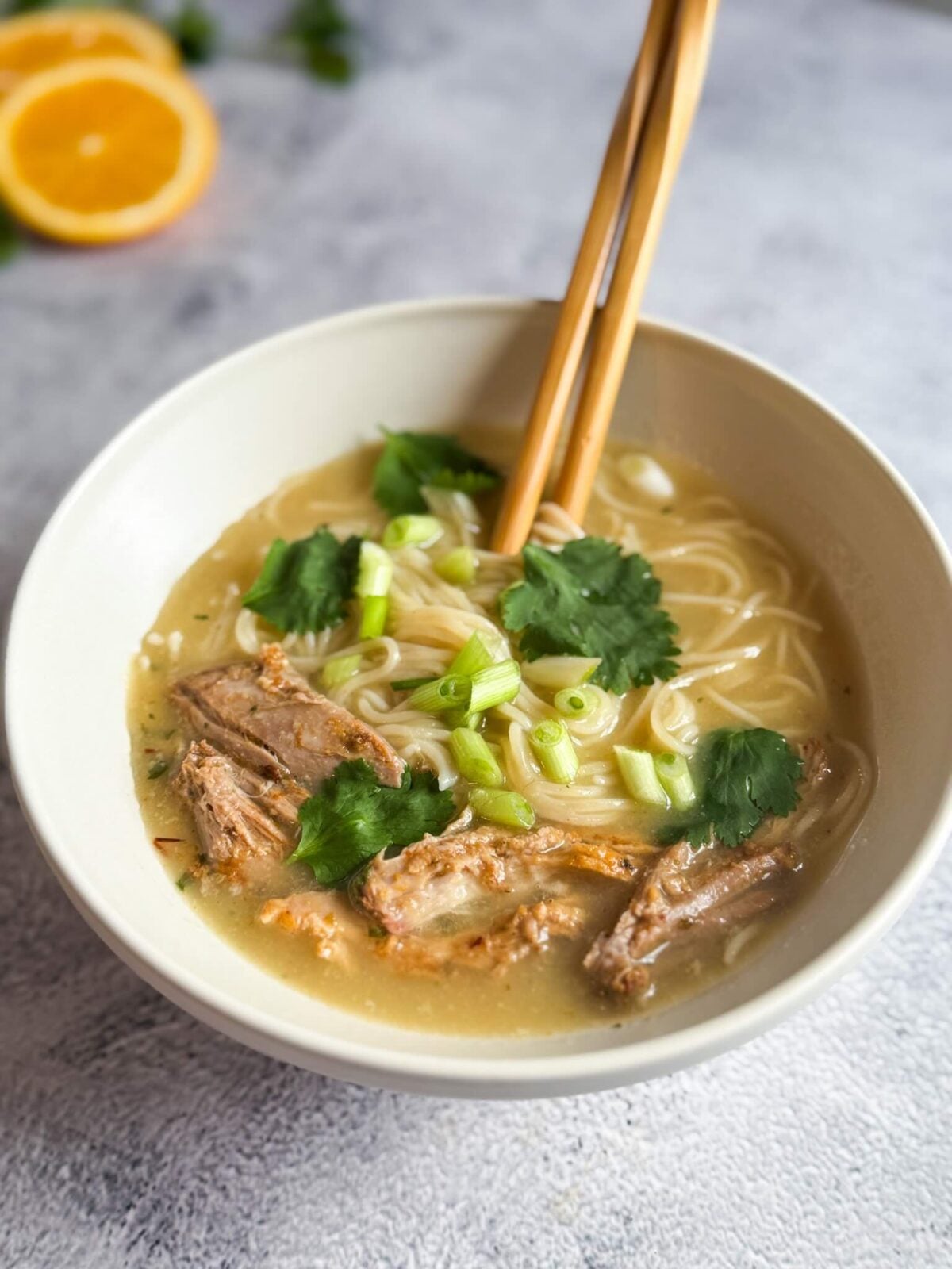 a white bowl of ramen with pork, cilantro, green onion, chopsticks, on gray backdrop.