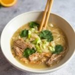 a white bowl of ramen with pork, cilantro, green onion, chopsticks, on gray backdrop.
